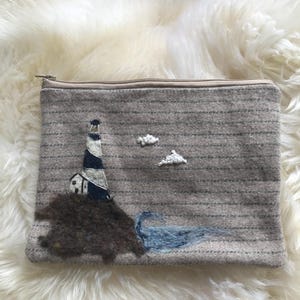 May include: A rectangular, gray fabric pouch with a zipper closure. The pouch features an embroidered coastal scene with a lighthouse, small house, two white clouds, and blue water. The lighthouse has black and white stripes. The pouch is on a white, fluffy surface.