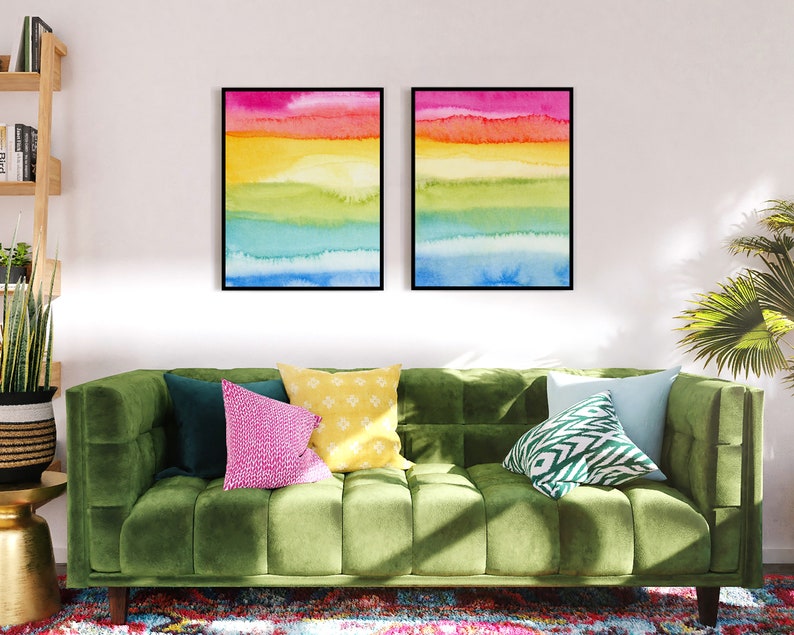 May include: Two framed watercolour paintings with a rainbow colour scheme. The paintings are hung on a white wall above a green velvet sofa with colourful throw pillows.