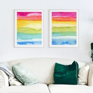 May include: Two framed watercolour paintings with a rainbow colour scheme. The paintings are hung on a white wall above a white sofa with green and dark green throw cushions.