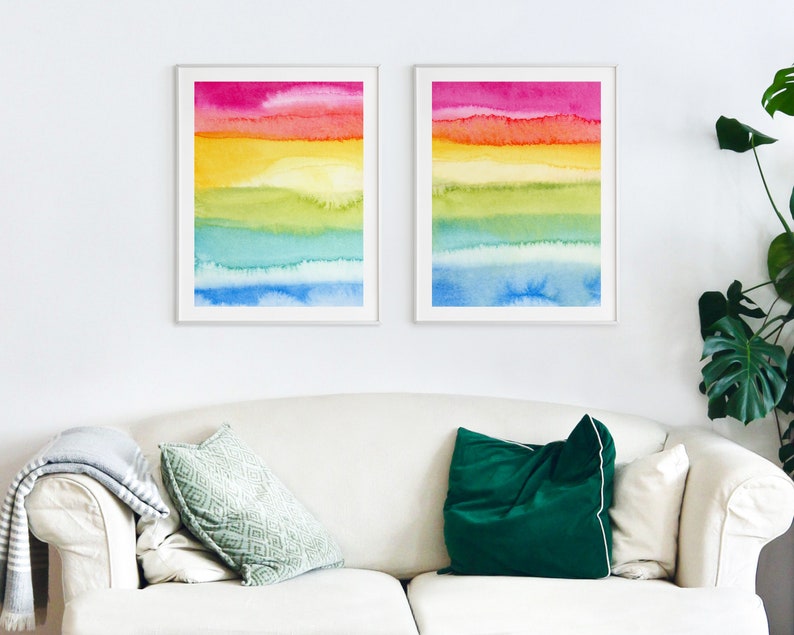 May include: Two framed watercolour paintings with a rainbow colour scheme. The paintings are hung on a white wall above a white sofa with green and dark green throw cushions.