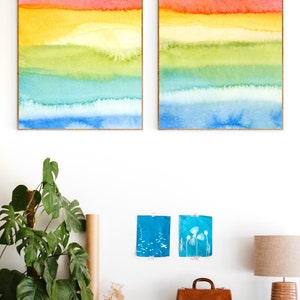 May include: Two framed watercolour paintings with horizontal stripes of pink, orange, yellow, green, blue, and light blue. The paintings are hanging on a white wall above a wooden dresser with a potted plant, a brown leather bag, and a camera.