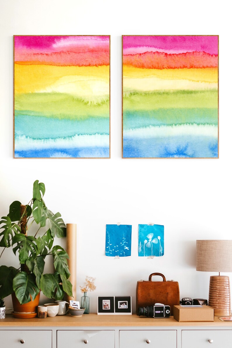 May include: Two framed watercolour paintings with horizontal stripes of pink, orange, yellow, green, blue, and light blue. The paintings are hanging on a white wall above a wooden dresser with a potted plant, a brown leather bag, and a camera.