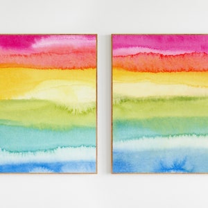 May include: Two framed abstract watercolour paintings with horizontal stripes of pink, orange, yellow, green, blue, and light blue.