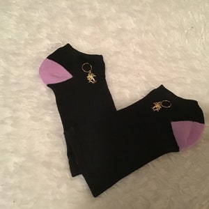 May include: A pair of black ankle socks with light purple heels and a gold-colored charm. Each sock has a gold-colored charm attached to the top. The socks are displayed on a white, fluffy surface.