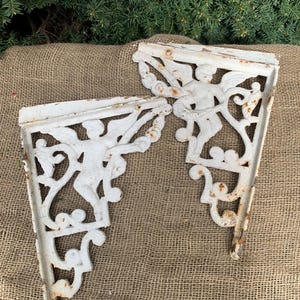 Può includere: Due staffe per mensole in ghisa bianca, ornate e con finitura anticata. Ogni staffa presenta un motivo decorativo a forma di angelo e volute. Le staffe sono esposte su una superficie di juta, suggerendo un'estetica vintage.