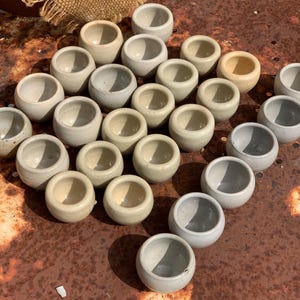 Op de afbeelding: Een verzameling kleine, ronde keramische kommen in verschillende tinten beige en grijs. De kommen zijn in een verspringend patroon op een roestig metalen oppervlak gerangschikt.