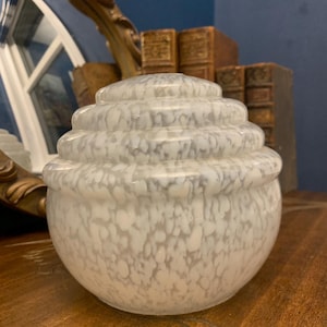 May include: A white and gray speckled glass jar with a tiered lid. The jar has a rounded base and a series of stacked, circular tiers leading up to the lid. The jar is sitting on a wooden surface.