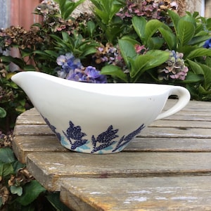May include: White ceramic gravy boat with a blue and white floral design. The boat has a handle and is sitting on a wooden table.