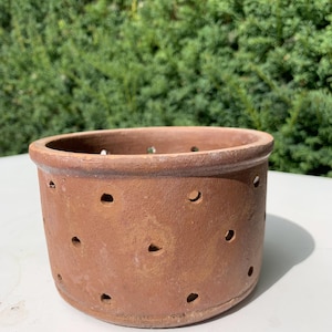 Pode incluir: Um vaso redondo de terracota sem vidrado com numerosos pequenos orifícios espaçados uniformemente. O vaso tem uma borda ligeiramente alargada e uma cor castanha quente e terrosa. Ideal para plantar ervas ou pequenas flores. O vaso está sobre uma superfície branca.