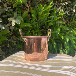 May include: A copper pot with two brass handles. The pot is cylindrical with a polished, reflective surface. The handles curve upwards. The pot sits on a striped, olive-green and white fabric, with a backdrop of green foliage.