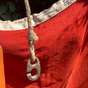 May include: A vibrant red fabric textile featuring a beige border with the number 602 printed in black. A thick, textured, light-coloured rope is attached to the fabric, ending in a metallic, C-shaped hook with a weathered, dark finish.
