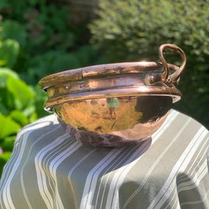 May include: A large copper bowl with a hammered finish, featuring a rounded base and a wide rim. It has a handle and sits on a striped tablecloth. The copper has a reddish-brown hue. The background shows blurred green foliage, typical of a garden setting.
