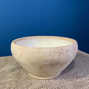 Può includere: Una piccola ciotola rotonda in terracotta con un esterno opaco color beige chiaro caratterizzato da naturali variazioni marrone terra e un interno con smalto bianco lucido. La ciotola presenta un design semplice con bordo largo.