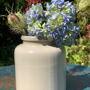 Puede incluir: Un jarrón de cerámica color crema contiene un ramo de hortensias azules y verdes, flores rojas y vainas de semillas. El jarrón tiene forma cilíndrica con una base ligeramente redondeada. El arreglo está sobre una superficie desgastada de color verde azulado.