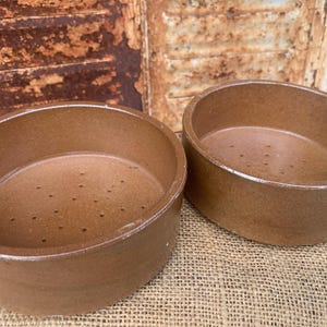 May include: Two round, brown ceramic bowls with smooth, glazed interiors and exteriors. Each bowl features a flat base with a series of small, evenly spaced drainage holes arranged in a circular pattern across the bottom surface of the vessel.
