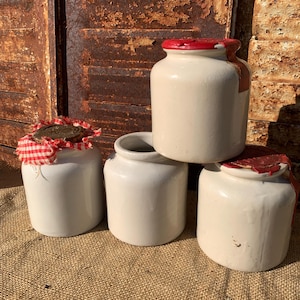 Op de afbeelding: Vier cilindervormige keramische potten met een gladde, gebroken witte afwerking. Eén pot is open, terwijl de andere zijn verzegeld met rode was of een rood-wit geblokte stoffen hoes die is vastgezet met een donkere waszegel.