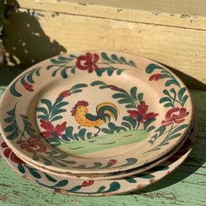 May include: Three antique ceramic plates stacked on top of each other. Each plate is decorated with a hand-painted design of a yellow rooster, red flowers, and green foliage. The plates have a cream background and a rustic, aged look.
