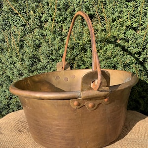 May include: An antique brass bucket with a curved handle. The bucket has a rounded shape with a wide rim and is supported by a burlap surface. The handle is made of a darker material, possibly leather, and is attached with rivets.