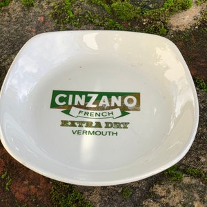 Op de afbeelding: Witte keramische asbak met een groen en gouden logo met de tekst "Cinzano French Extra Dry Vermouth".