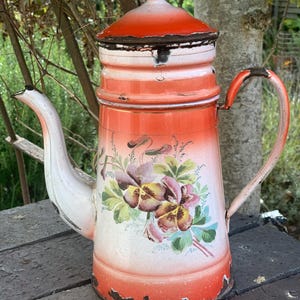 May include: A vintage enamel coffee pot with a white spout and handle, and a red and white body. The pot features a floral design with purple, yellow, and green flowers. The pot shows signs of wear, adding to its antique charm.