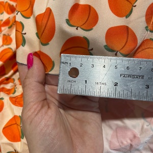 May include: Peach print fabric with a ruler measuring 2 inches. The fabric is a light cream color with orange peaches printed on it.