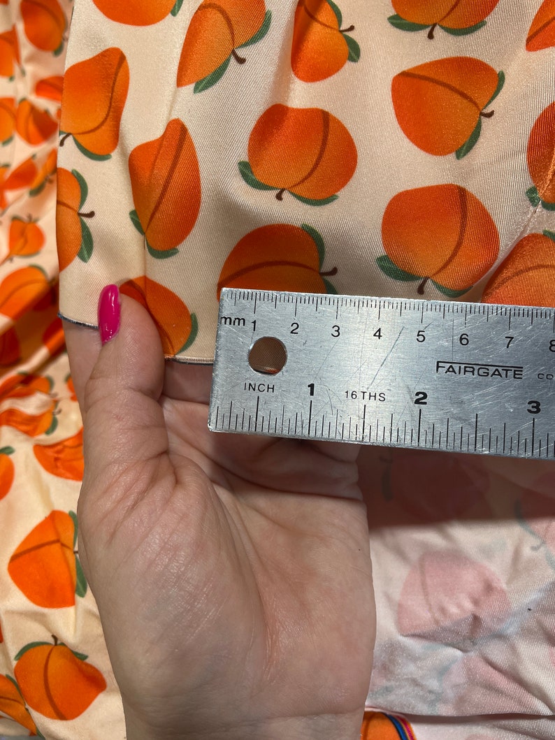 May include: Peach print fabric with a ruler measuring 2 inches. The fabric is a light cream color with orange peaches printed on it.