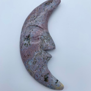Può includere: Una scultura in pietra a forma di luna crescente con un viso scolpito al suo interno. La pietra è di colore viola chiaro con macchie più scure di viola e bianco.