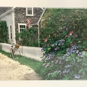 May include: A scenic photograph of a coastal home with a shingled roof and white picket fence. A bicycle with a basket is parked near the fence. An American flag waves in the breeze. Lush green and purple flowering bushes are in the foreground.