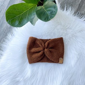 Peut inclure: Un bandeau en tricot marron avec un d&eacute;tail nou&eacute;. Le bandeau est sur une surface blanche et moelleuse.