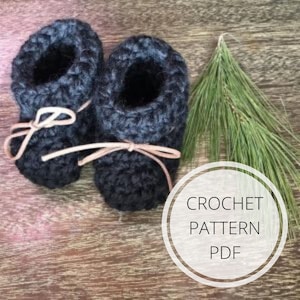 Peut inclure: Une paire de chaussons pour bébé en crochet gris foncé avec des liens en cuir marron. Les chaussons sont sur une surface en bois avec une branche de pin et un cercle blanc avec le texte "CROCHET PATTERN PDF".