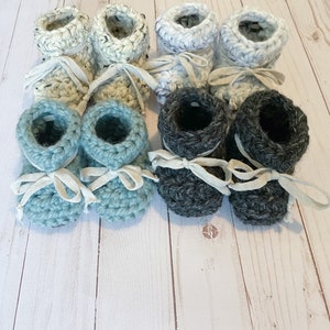 May include: Six pairs of crocheted baby booties in blue, grey, and white. The booties have a simple design and are tied with white ribbon.