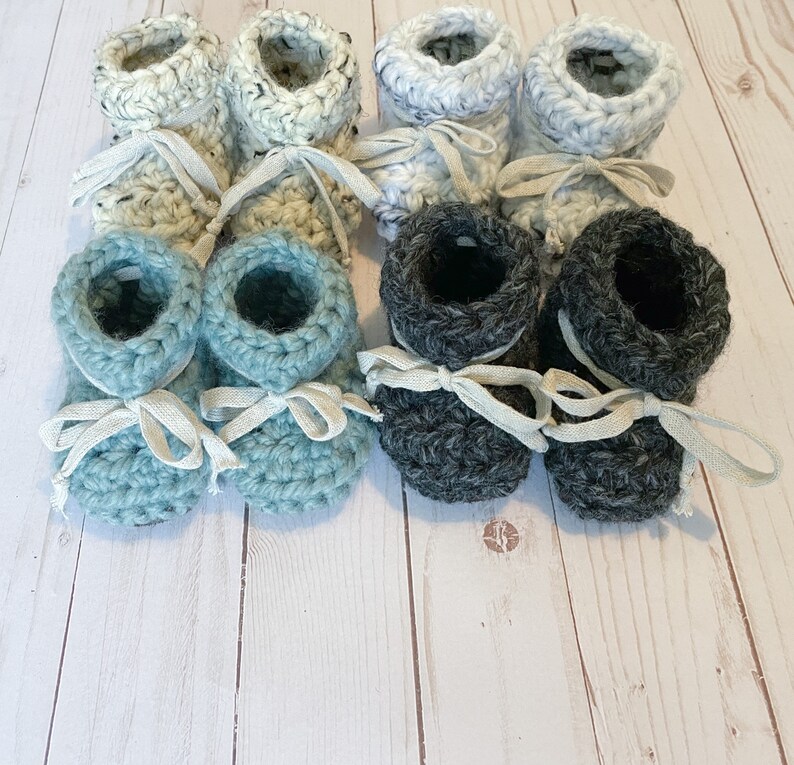 May include: Six pairs of crocheted baby booties in blue, grey, and white. The booties have a simple design and are tied with white ribbon.
