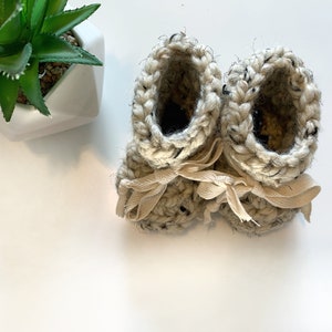 May include: A pair of cream-coloured crocheted baby booties with black flecks and cream-coloured ribbon ties.
