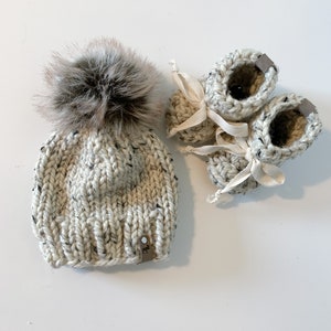 May include: A white knitted baby hat with a brown faux fur pom pom and matching booties. The booties have white ribbon ties.