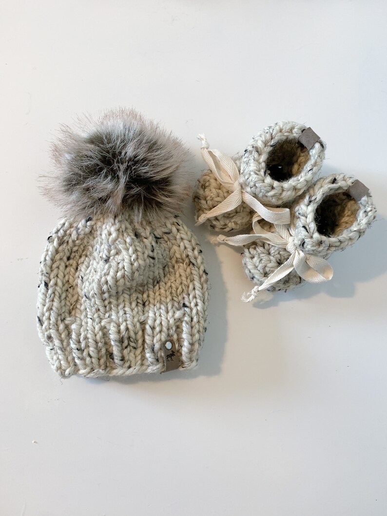 May include: A white knitted baby hat with a brown faux fur pom pom and matching booties. The booties have white ribbon ties.