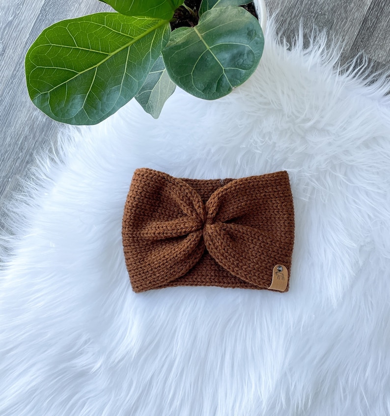 Peut inclure: Un bandeau en tricot marron avec un d&eacute;tail nou&eacute;. Le bandeau est sur une surface blanche et moelleuse.