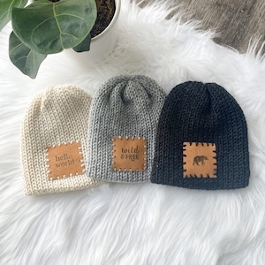 May include: Three knitted beanies with leather patches. The beanies are in beige, gray, and black. The patches have the text "hello world", "wild & free", and a bear silhouette.