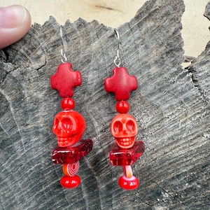 May include: Red skull earrings with silver hooks. Each earring features a red cross, a skull, and a decorative base with a swirl design. The earrings are displayed on a textured wooden surface.