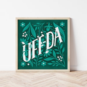 May include: A teal green framed print with white lettering that says "UFF DA" surrounded by white flowers and green leaves.