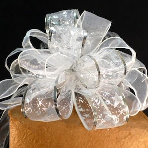 May include: A large, decorative bow made of sheer white ribbon with silver accents and a subtle snowflake pattern. The bow is atop a brown, textured surface, creating a festive and elegant look. Perfect for gift wrapping or holiday decorations.