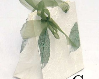 May include: A white paper gift bag with a green leaf design and a green ribbon tied around the handles. The bag is labeled with the letter "G".