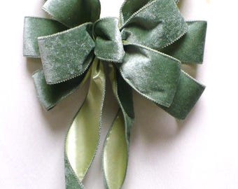 May include: A sage green velvet bow with a satin center and long, flowing tails. The bow is tied in a classic style, with multiple loops and a central knot. The edges of the velvet are finished with a contrasting trim.