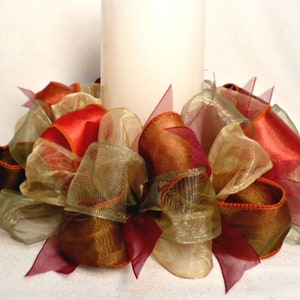 May include: A white candle is surrounded by a wreath of brown, gold, and burgundy sheer ribbon. The ribbon is arranged in a decorative, layered pattern.