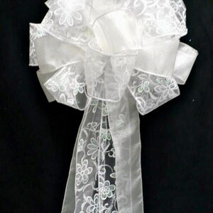 May include: A large, decorative bow made of sheer white ribbon with a delicate embroidered floral pattern. The ribbon features a translucent texture and a repeating design of small flowers and swirling vines, with two long tails hanging down from the center.