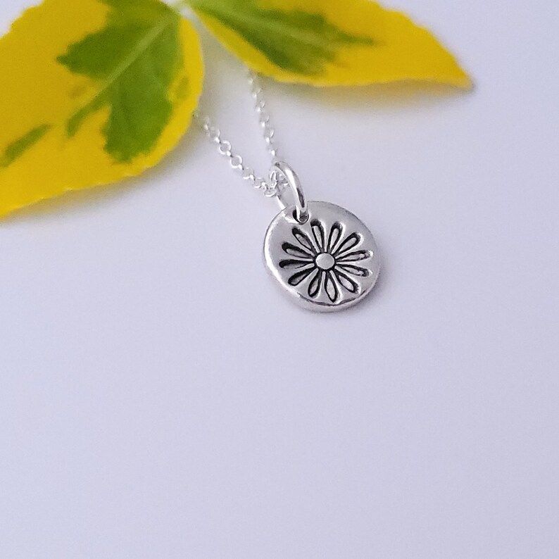 May include: A silver necklace with a small, round pendant featuring a daisy design. The pendant is textured and has a slightly raised center.