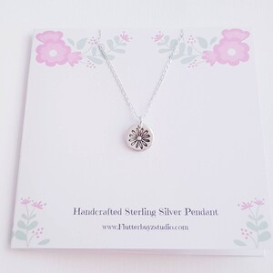 May include: A silver necklace with a small, round pendant featuring a daisy design. The necklace is displayed on a white card with a floral pattern and the text "Handcrafted Sterling Silver Pendant" and "www.Flutterbuyzstudio.com".