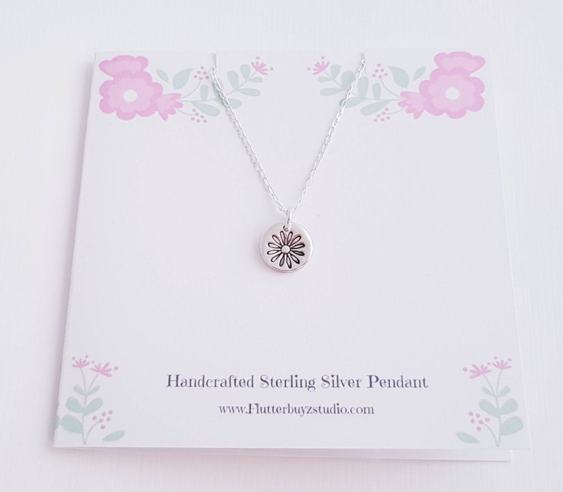 May include: A silver necklace with a small, round pendant featuring a daisy design. The necklace is displayed on a white card with a floral pattern and the text "Handcrafted Sterling Silver Pendant" and "www.Flutterbuyzstudio.com".