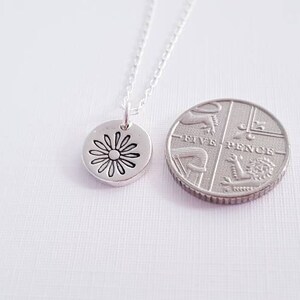 May include: A silver necklace with a small, round pendant featuring a daisy design. The pendant is shown next to a British five pence coin for size comparison.
