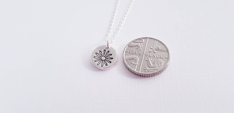 May include: A silver necklace with a small, round pendant featuring a daisy design. The pendant is shown next to a British five pence coin for size comparison.