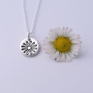 May include: A silver necklace with a small, round pendant featuring a daisy design. The pendant is textured and has a raised center. A white daisy with a yellow center is shown next to the necklace.